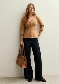 Mujer vistiendo un cárdigan color camello, jeans oscuros de pierna ancha, zapatos negros, sosteniendo un bolso de cuero marrón, de pie contra una pared clara.