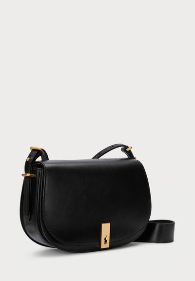 Sac en cuir noir en bandoulière avec une forme incurvée, des finitions en métal doré et une fermeture à rabat avec une plaque logo rectangulaire.