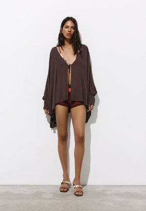 Femme portant un cardigan ample marron foncé par-dessus un haut de bikini à motifs et un short de bain bordeaux, debout contre un mur blanc avec des sandales aux pieds.