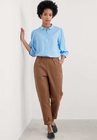 Svetlo modra srajca z naborki in napihnjenimi rokavi, v kombinaciji z rjavimi hlačami visoke oblazitve. Model nosi črne sandalke brez petke pred nevtralno ozadje.