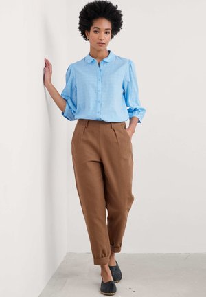 Camicia azzurro chiaro con maniche a sbuffo, abbinata a pantaloni marroni a vita alta. Modella che indossa scarpe nere senza lacci su uno sfondo neutro.