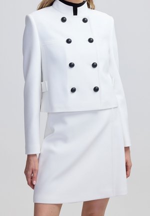 Biała, dopasowana marynarka z sześcioma czarnymi guzikami, zestawiona z pasującą białą spódnicą, noszona przez osobę na jednolitym tle.