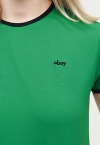 Πράσινο μπλουζάκι με μαύρο γείσο στο γιακά και στα μανίκια, με μικρό μαύρο κεντημένο κείμενο "obey" στο αριστερό μέρος του στήθους.