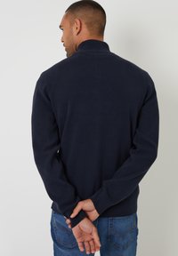 Maglione a maglia blu navy con alto collo, maniche lunghe e polsini a coste. Indossato con jeans di denim, mostrando una vista posteriore semplice.