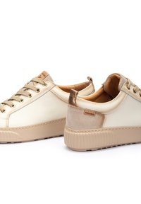 Zapatillas de cuero crema con acentos topo, que cuentan con una suela de goma texturizada, ojales dorados y una pequeña etiqueta con el logo en el talón.