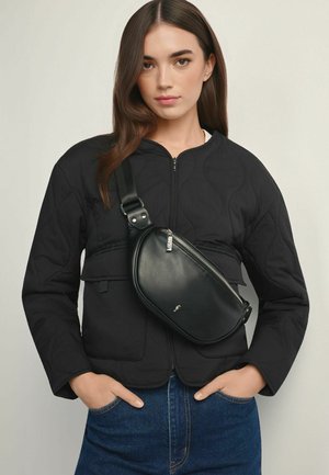 Młoda kobieta z długimi brązowymi włosami, ubrana w czarną pikowaną kurtkę, niebieskie dżinsy oraz czarną skórzaną torebkę na ramię z zamkiem błyskawicznym.