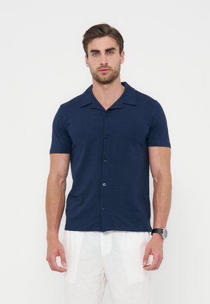 Hombre con camisa azul marino de manga corta con botones y pantalones blancos, de pie frente a un fondo blanco liso, mirando hacia adelante.