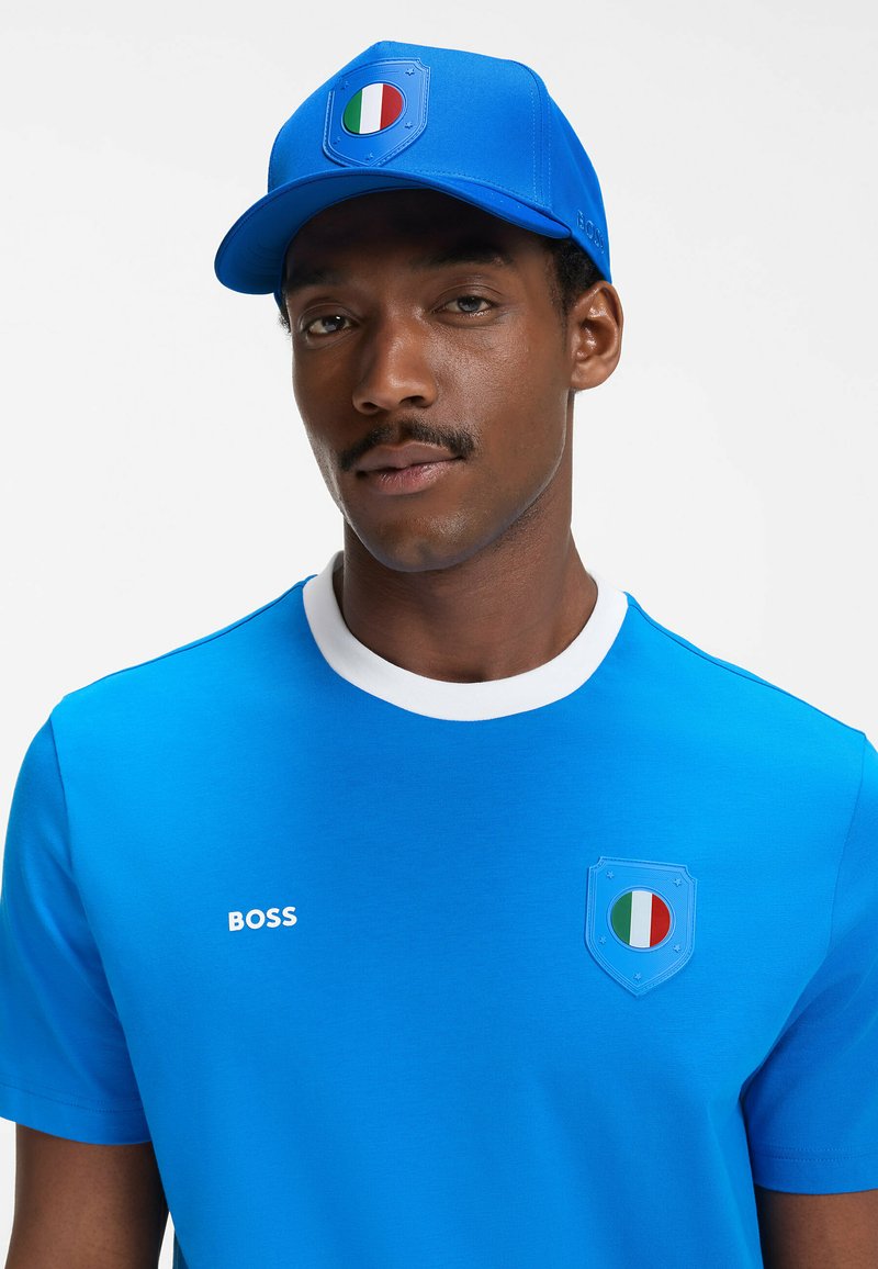 Homme portant une casquette bleue et un T-shirt avec des emblèmes du drapeau italien et le logo "BOSS" sur la poitrine, sur un fond uni.