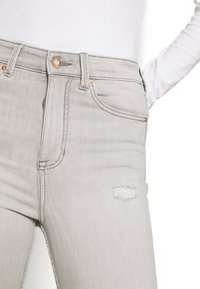 Jeans de denim gris claro con corte ajustado, que cuentan con bolsillos delanteros, un pequeño parche desgastado y herrajes tono cobre en la cinturilla.