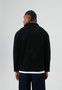 Schwarze, flauschige, strukturierte Jacke mit Kragen und langen Ärmeln, kombiniert mit einem weißen Hemd und dunkler Jeanshose. Ansicht von hinten.