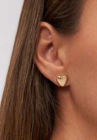 Primo piano di un orecchio con capelli castani lisci e dritti e un piccolo orecchino a forma di cuore, lucido e dorato.
