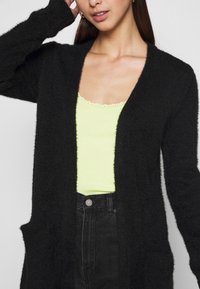 Kvinna iklädd en gul toppen med spetskant, svart cardigan och högmidjade svarta jeans, med handen som rör vid sitt huvud.