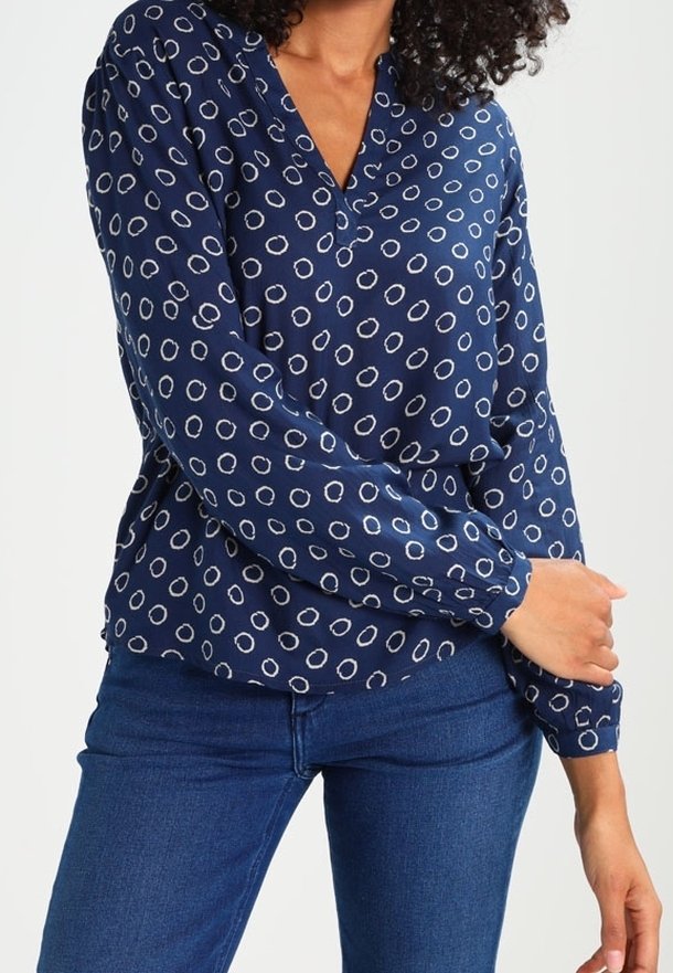 Blouse marine à motif circulaire blanc, col en V, manches longues et texture douce. Associée à un jean bleu.