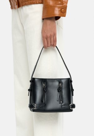 Ręka trzyma małą czarną skórzaną torebkę z dwoma przednimi suwakami i srebrnymi klamrami, ubrana w białe spodnie i brązową skórzaną kurtkę.