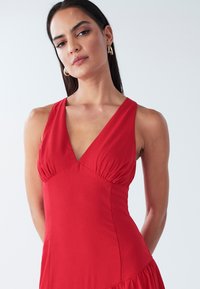 Red sleeveless dress with a deep V-neck, gathered fabric at the bust, and a flared hemline. Made from smooth fabric with a simple design.