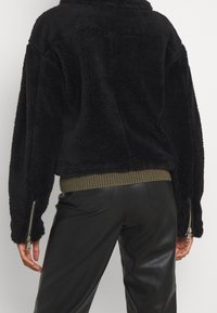 Sort fake pelsjakke med en cropped design, olivengrøn ribbestrikket kant og lynlås detaljer på ærmerne; har en tekstureret, blød overflade.