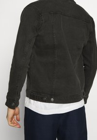 Donkerdenimjack met een klassiek ontwerp, knoopaccenten en zichtbare stiksels. Draagt over een wit shirt, met een gestructureerde stof.