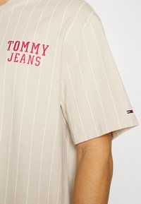 Béžové tričko s krátkým rukávem a bílými vertikálními pruhy. Červená výšivka "TOMMY JEANS" na hrudi. Na rukávu je malý akcent v podobě vlajky.
