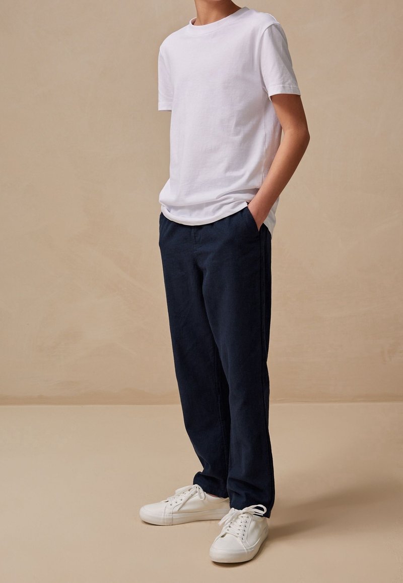 Personne portant un t-shirt blanc à manches courtes uni, un pantalon bleu marine avec les mains dans les poches, et des baskets blanches à lacets, sur fond beige.