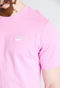 Vaaleanpunainen puuvillapaita, jossa on valkoinen brodeerattu Nike-logo vasemmassa rinnassa. Pyöreäkauluksinen malli, jossa on lyhyet hihat ja sileä pinta.