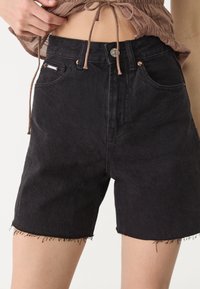 Mustat farkkushortsit, joissa on korkea vyötärö, revitty helma, kaksi etutaskua ja pieni metallinen nappisuljenta. Teksturoitu kangas, istuva malli.