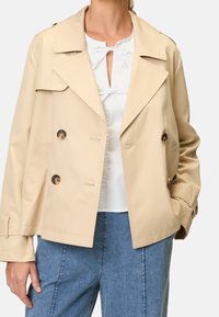 Beige dobbelbrystet trenchjakke med store klapper, to frontlommer og skinnende brune knapper. Letvægtstof med en glat tekstur.