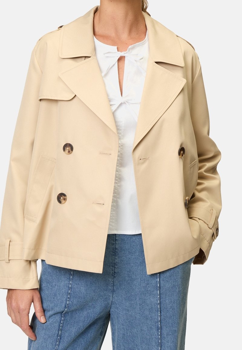 Beige dobbelbrystet trenchjakke med store klapper, to frontlommer og skinnende brune knapper. Letvægtstof med en glat tekstur.