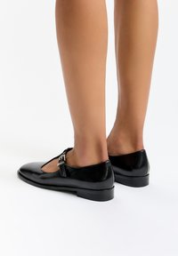 Musta nahkainen Mary Jane -kenkä pyöreällä varvaskärjellä, matalalla kantapäällä ja sivulla soljella. Sileä pinta ja klassinen muotoilu.