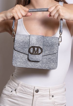 Vrouw in wit topje houdt gestructureerde grijze handtas met metalen sluitingslogo en afneembare band op borsthoogte vast.