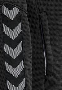 Tejido atlético negro con un patrón texturizado, que presenta un panel lateral con rayas en chevrón en blanco y negro. Incluye dos bolsillos.