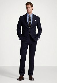 Tamsiai mėlynas kostiumas su pritaikytu pjūviu, derinamas su šviesiai mėlynu languotu marškinėliu, dryžuota kaklaraiščiu ir rudais odiniais batais. Yra balta kišenėlė.