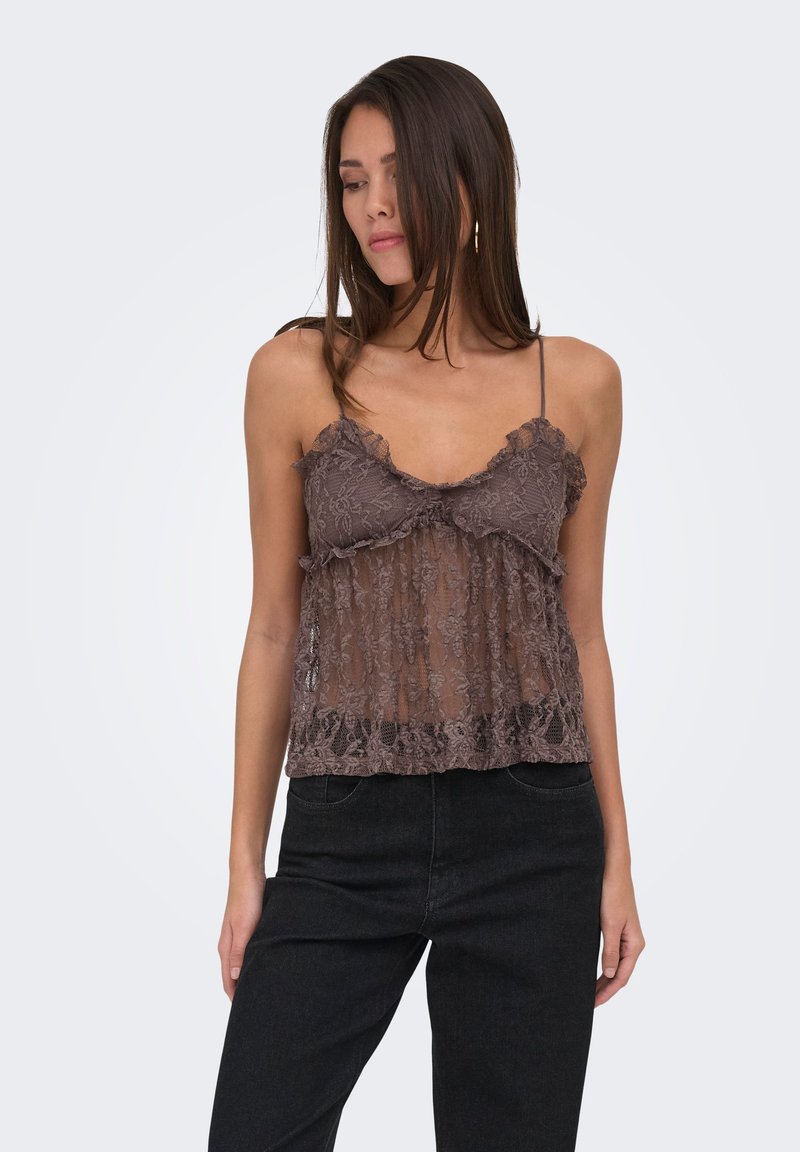 Femme portant un haut marron en dentelle sans manches avec de fines bretelles et un pantalon noir, regardant vers le bas sur un fond clair uni.