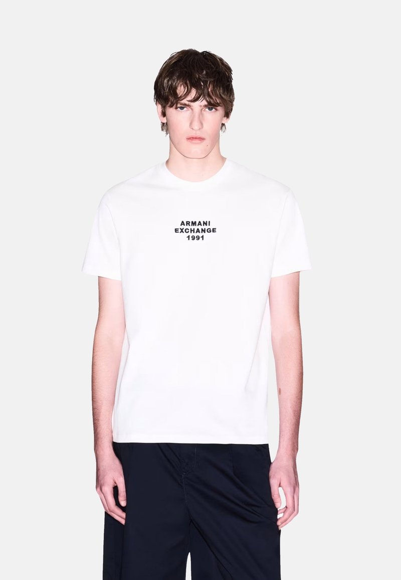 Valkoinen puuvillapaita, jossa on musta painettu logo "ARMANI EXCHANGE 1991" keskellä rintaa. Pyöreä pääntie, lyhyet hihat, rento malli.