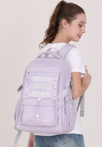 Sac à dos lavande avec plusieurs compartiments, accents en maille, bretelles réglables et poignée supérieure. Le matériau semble durable et texturé.