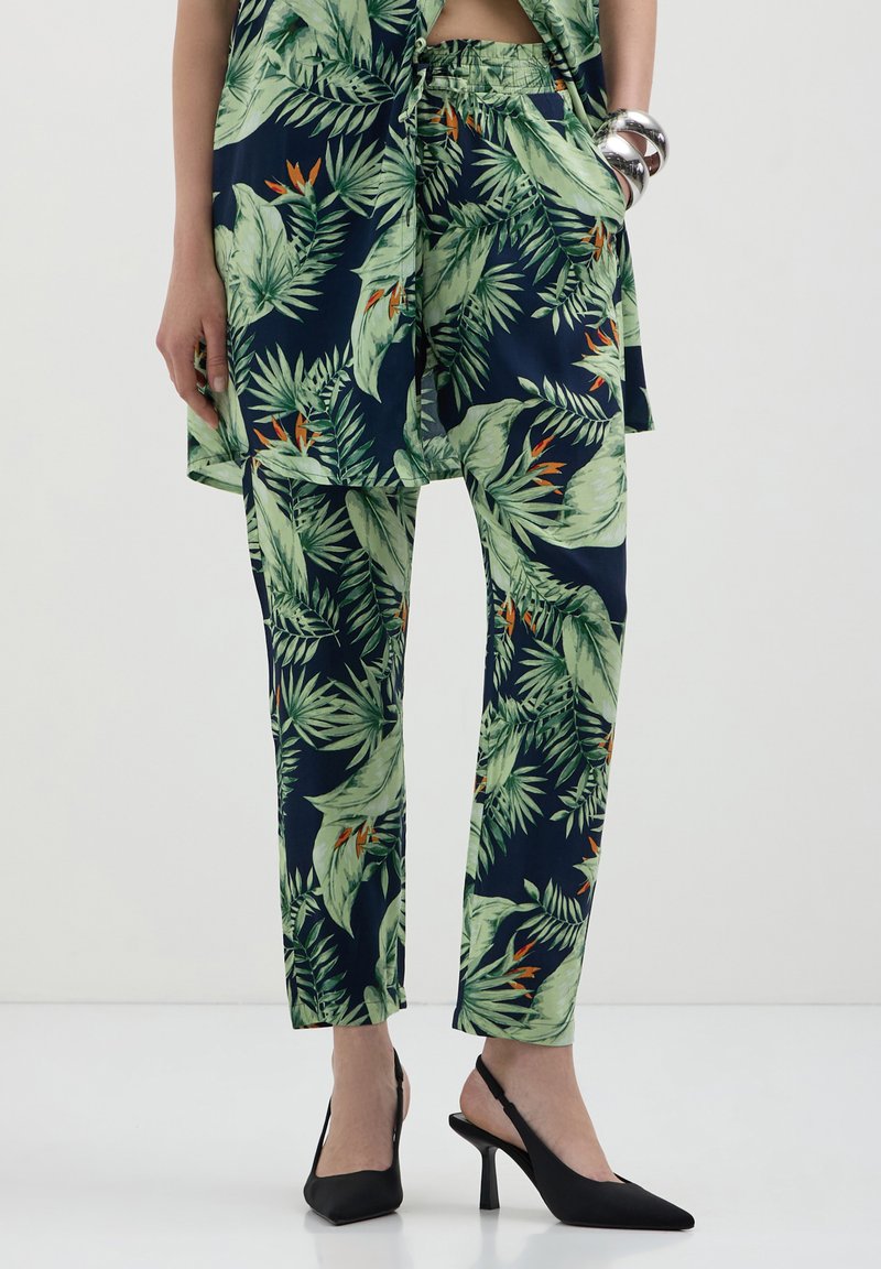 Tenue à motif tropical avec des feuilles vertes et des accents orange sur un fond bleu foncé ; haut léger et pantalon fuselé, associés à des talons noirs.