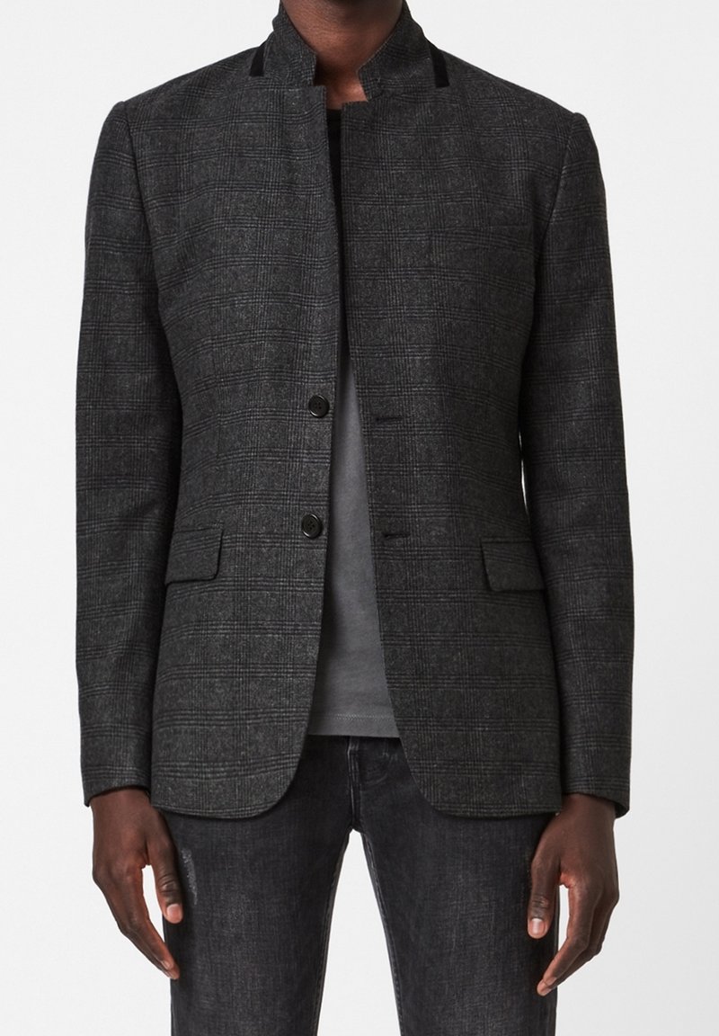 Šedý kostkovaný blazer vyrobený z texturovaného materiálu, s stojáčkovým límcem, dvěma předními kapsami a černými knoflíkovými zapínáními. Noste přes šedý top.