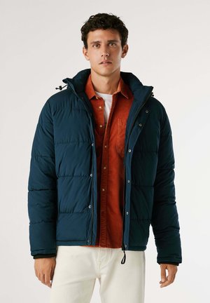 Chaqueta acolchada en verde azulado oscuro con capucha, que presenta un cierre frontal con cremallera, botones a presión y un diseño acolchado. Lleva una camisa naranja de botones debajo.