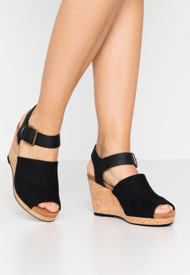 Jambes portant des sandales compensées noires à bout ouvert avec semelles en liège et brides réglables à la cheville sur un fond clair uni.