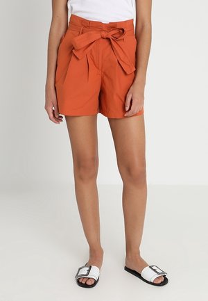 Woman wearing high-waisted burnt orange shorts with a front bow tie, paired with a white sleeveless top and white slide sandals with buckles.