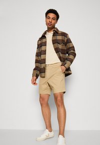 Veste en laine à carreaux marron et bleu, polo beige, shorts kaki et baskets blanches. La tenue présente des textures superposées et des tons neutres.