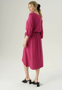 Abito fucsia a metà lunghezza con vita regolabile, maniche a tre quarti e texture liscia. Indossato con sandali neri con tacco bloccato.