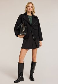 Zwarte bomberjack met rits, twee voorzakken, gecombineerd met een geplooide zwarte rok en kniehoog glanzende zwarte laarzen. Zwarte handtas in de hand.