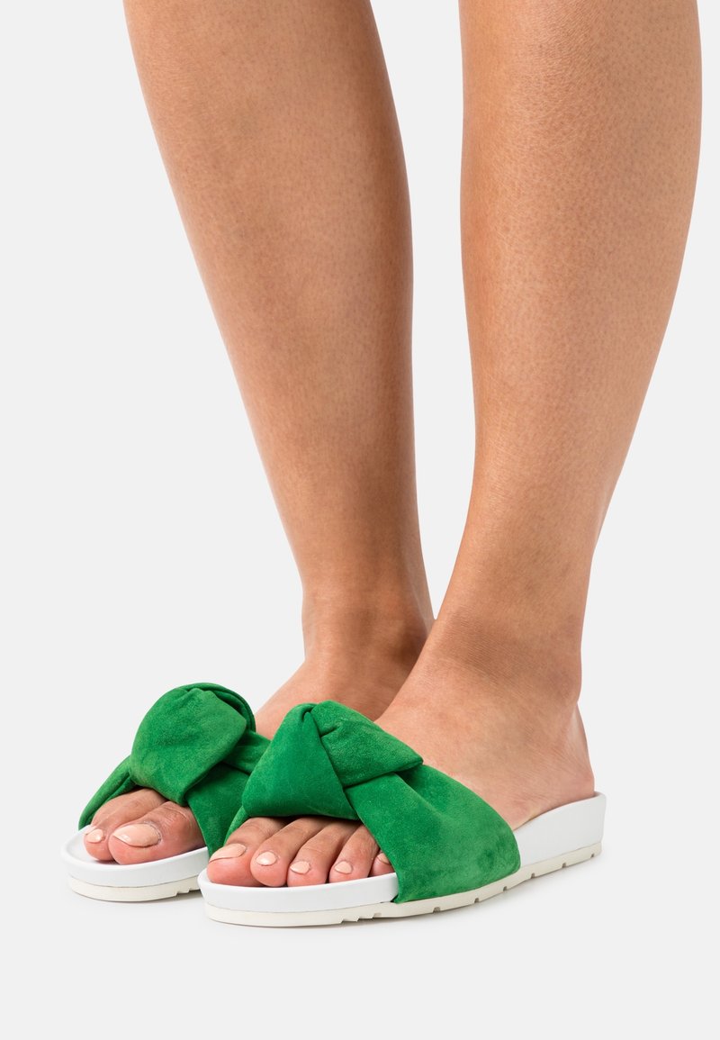 Sandales à glissière en daim vert avec un design de nœud, semelle en caoutchouc blanc et orteils découverts, reposant sur des pieds nus contre un fond neutre.