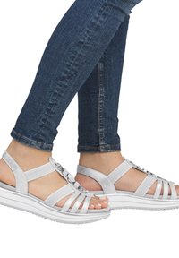 Sandali d'argento con un design a-strisce, dotati di due accenti circolari. Hanno una suola piatta bianca e sono abbinati a jeans in denim blu.
