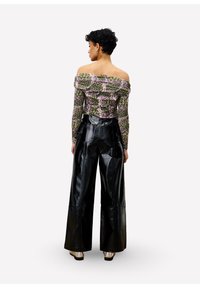 Vrouw met kort, krullend haar, gekleed in een off-shoulder gebloemd top en hoge, zwarte, wijde leren broeken, staand met haar rug naar de camera.