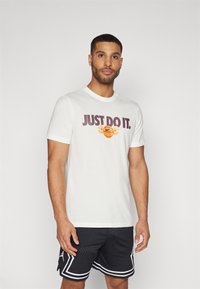 Hvit bomull T-skjorte med "JUST DO IT" i mørk lilla tekst og en basketballgrafikk med oransje aksenter. Brukt med svarte treningsshorts.