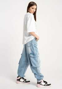 Kobieta ubrana w oversize'ową białą koszulkę, jasnoniebieskie spodnie cargo i czarno-biało-różowe sneakersy, stojąca z jedną ręką w kieszeni.