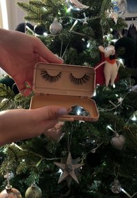 Falska ögonfransar i ett beige fodral, som innehåller två par med varierande längder. Inkluderar en liten spegel och är placerad framför en julgran.