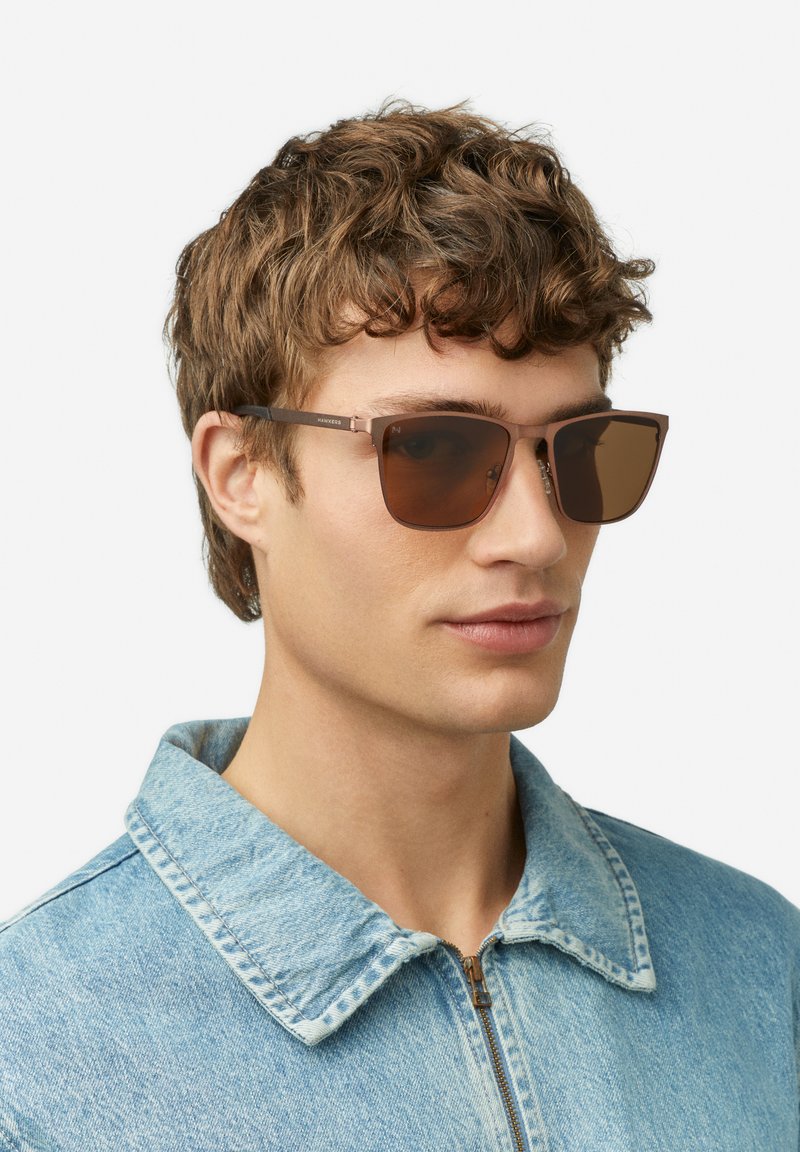Lunettes de soleil marron au design carré avec des accents métalliques, posées sur une veste en jean claire à col replié.