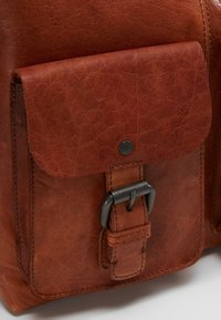 Bruine leren tas met een voorflapzak, metalen drukknoopsluiting en gestikte details. De textuur is soepel met een licht versleten uitstraling.